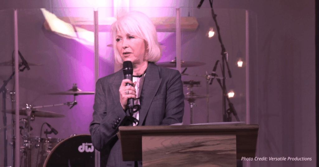 Tina Peters speaks at a podium in front of a church stage lit in purple, with a cross illuminated behind her.