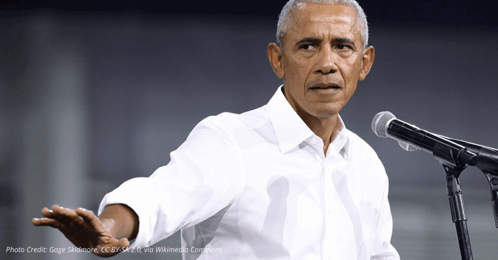 Barack Obama speaking at a campaign rally in a white shirt with hand extended near microphone