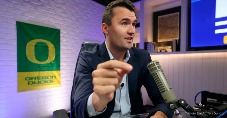 Charlie Kirk mid-gesture at a mic, Oregon Ducks flag on wall