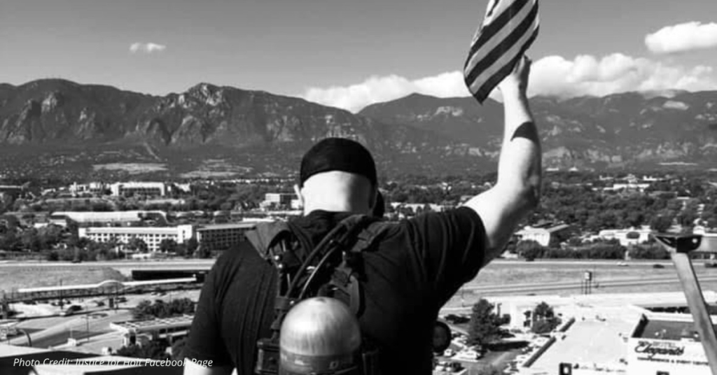Firefighter raising an American flag from a rooftop overlooking Colorado mountains