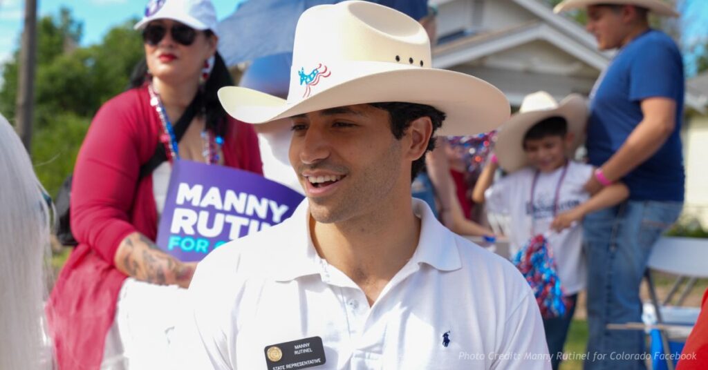 Manny Rutinel wearing a white cowboy hat at a community event while supporters hold a campaign sign behind him.