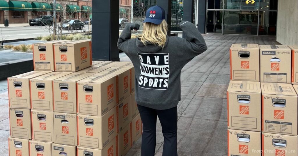 Volunteer stands between stacks of petition boxes outside a Denver building wearing a “Save Women’s Sports” sweatshirt.