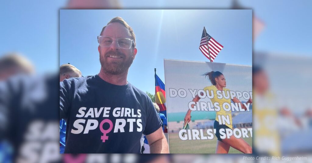 Rich Guggenheim at a rally wearing a “Save Girls Sports” shirt, standing in front of a sign asking “Do you support girls only in girls sports?”
