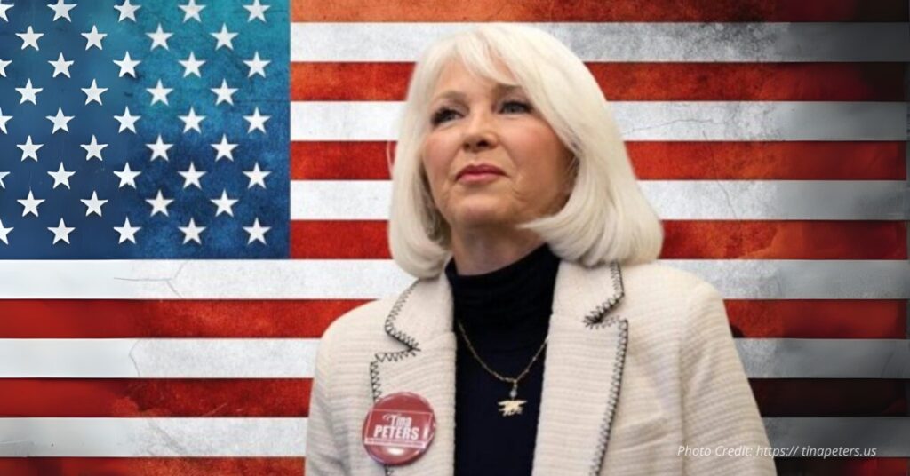 Tina Peters stands in front of an American flag backdrop wearing a Tina Peters campaign button