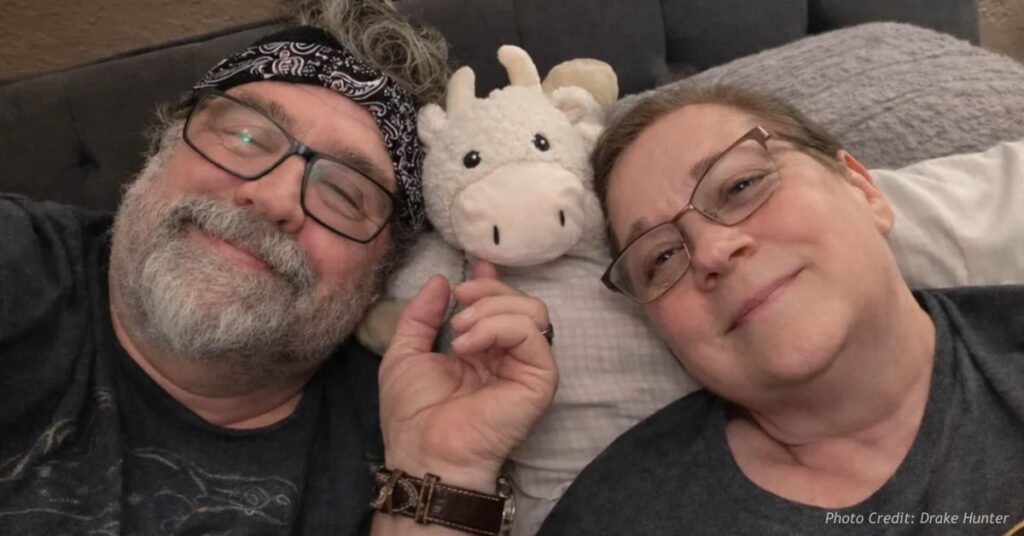 Drake and Sherrie Hunter lie side by side, heads together with a small stuffed cow between them, both smiling at the camera.