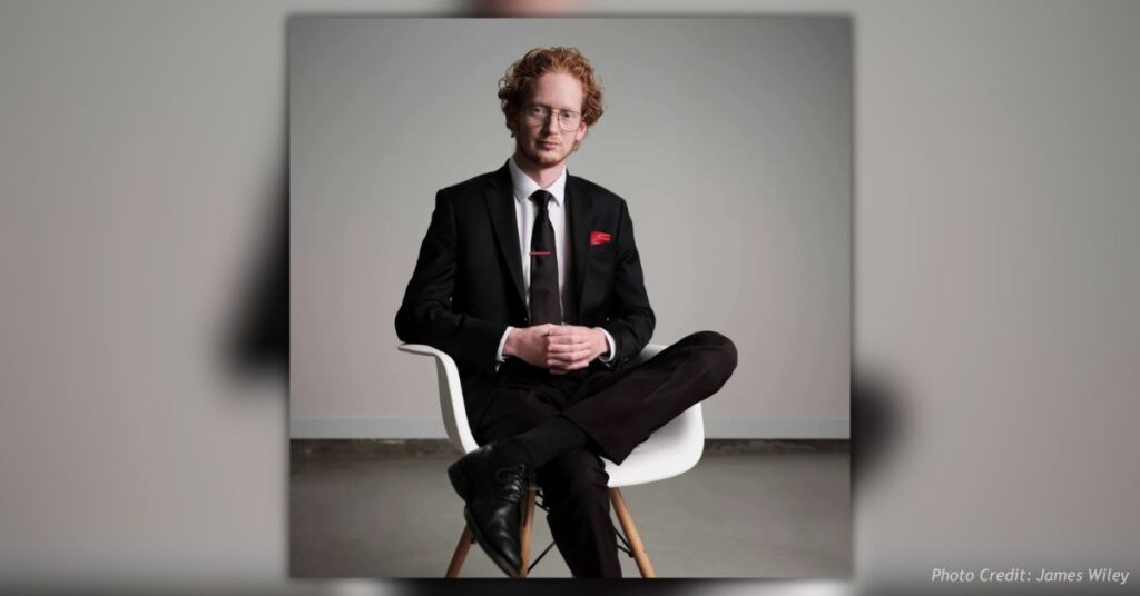 James Wiley, Colorado Secretary of State candidate, seated in a white chair wearing a black suit, black tie, glasses, and a red pocket square against a plain gray background